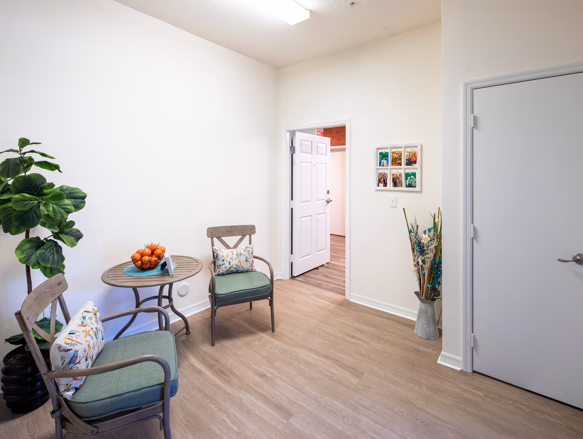 A small seating area with two wooden chairs featuring green cushions and patterned pillows, a round wooden table with a bowl of oranges, a tall green potted plant, a vase with decorative dried flowers, and two white doors in a room with light wood flooring and white walls.