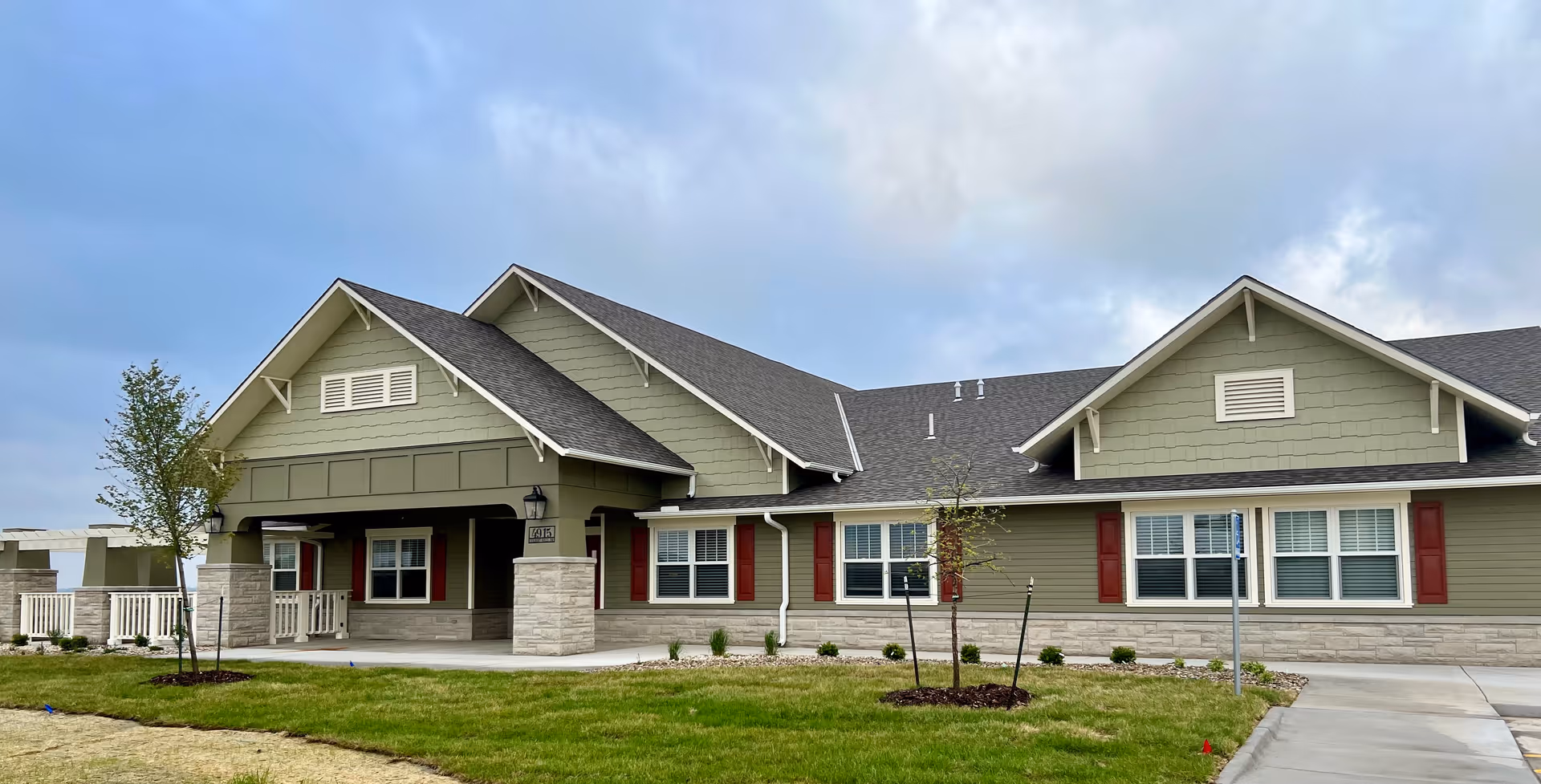 Single-story green and beige assisted living building with gabled roofs, a covered entrance, and a landscaped lawn under a cloudy sky.