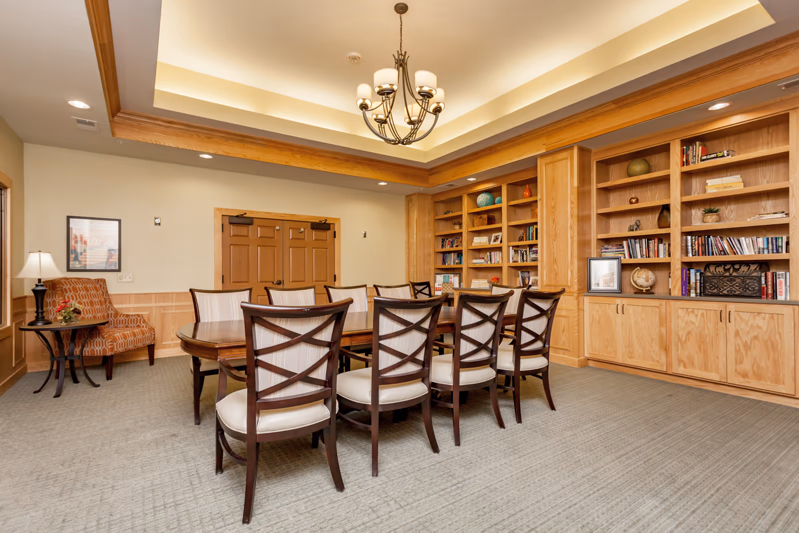 A well-lit room with a long wooden table surrounded by eight cushioned chairs. The room features built-in wooden bookshelves filled with books and decorative items along one wall. There is a patterned armchair with a small side table and lamp in the corner. The ceiling has recessed lighting and a chandelier with multiple lights.