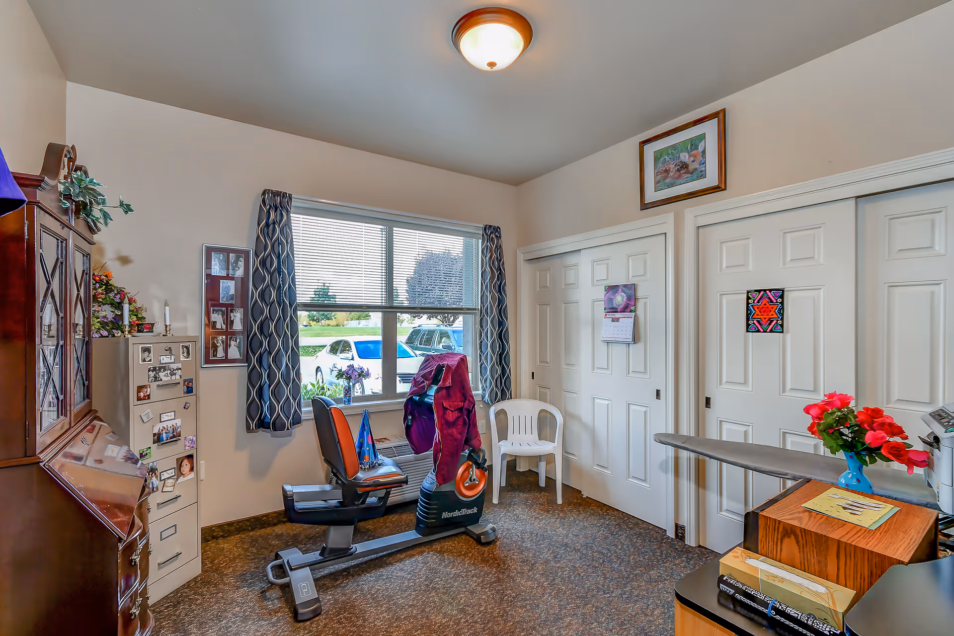 A small room with a recumbent exercise bike positioned near a window with patterned curtains. The room has a filing cabinet with photos and magnets, a wooden cabinet, a white plastic chair, and two closed white sliding closet doors with decorations on them. A framed picture hangs above the closet doors, and a small table with a vase of red flowers and some papers is visible in the foreground.