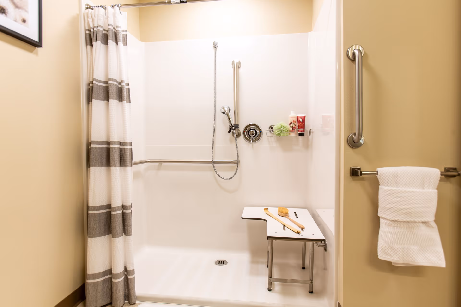 Accessible shower area with a white and gray striped shower curtain, a foldable shower seat with a wooden brush on it, grab bars on the wall, a handheld showerhead, and toiletries on a small shelf inside the shower.