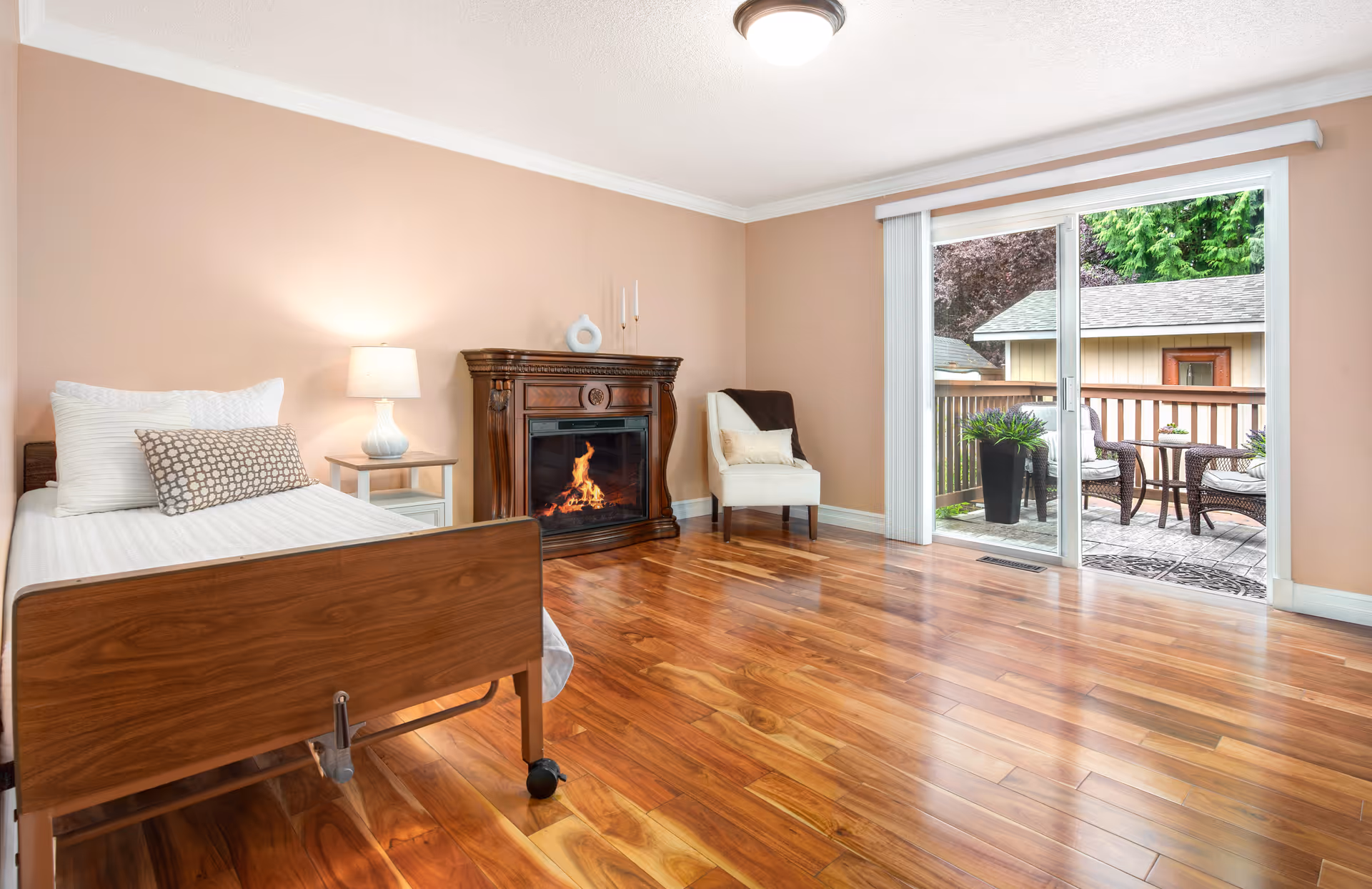 A cozy bedroom with a wooden bed featuring white bedding and pillows, a bedside table with a lamp, a decorative fireplace with a fire burning, a white armchair, and a sliding glass door leading to an outdoor patio with chairs and a table surrounded by greenery.