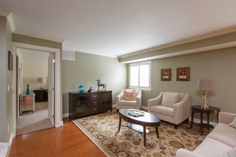 A cozy living room with light green walls, hardwood floors, and a patterned area rug. The room features a beige loveseat, two beige armchairs, a wooden coffee table with decorative items, a side table with a lamp, and a dark wooden cabinet with decorative pieces on top. A window with white shutters lets in natural light, and an open doorway leads to a bedroom with a nightstand and lamp visible.