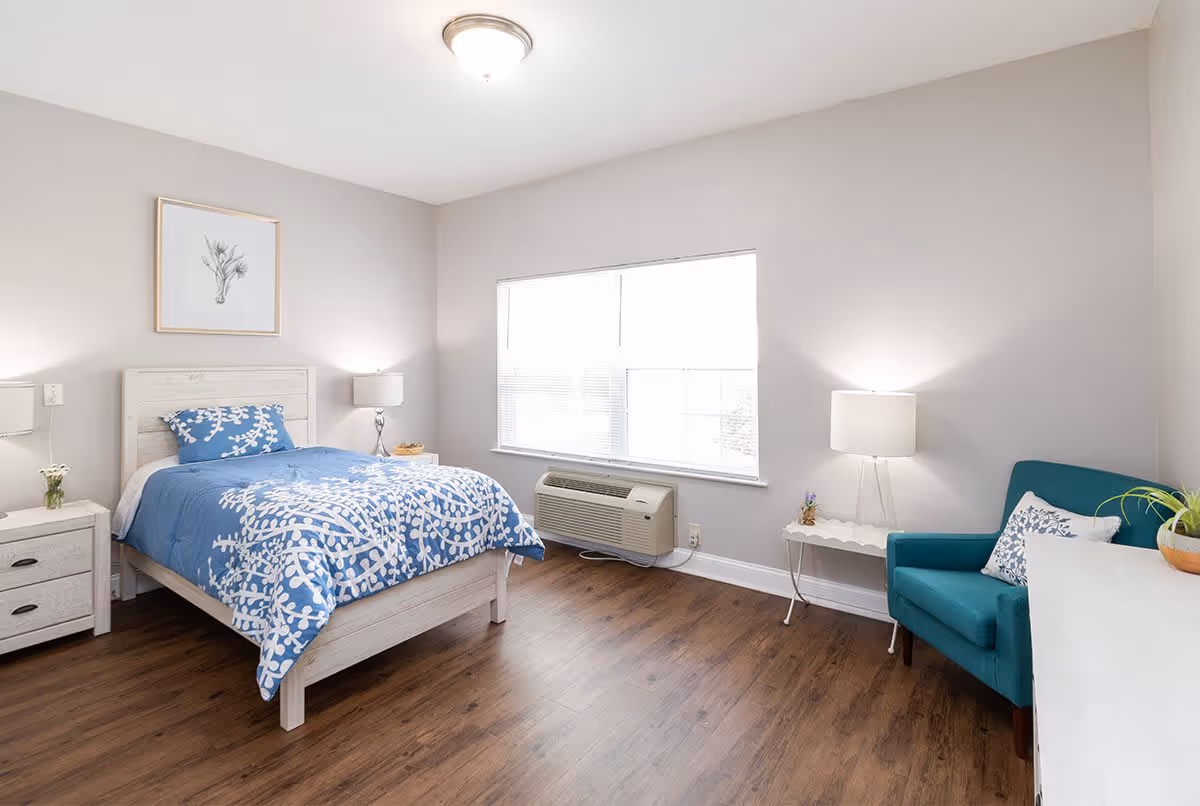 A bright and clean bedroom with a single bed featuring blue and white patterned bedding. The room has light gray walls, a wooden floor, a window with blinds, a white nightstand with a lamp and small plant, a teal armchair with a decorative pillow, and a white desk with a potted plant. A framed botanical print hangs above the bed.