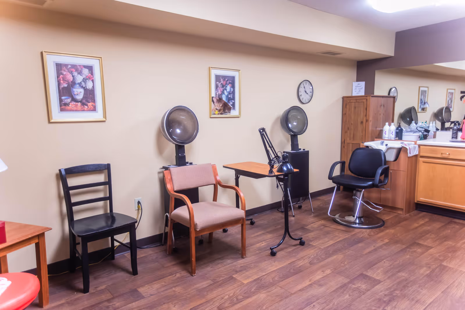 Interior view of a salon area in an assisted living facility with wooden flooring, two hair drying stations with chairs, a black salon chair, a wooden cabinet, a sink, and framed floral artwork on the beige walls.