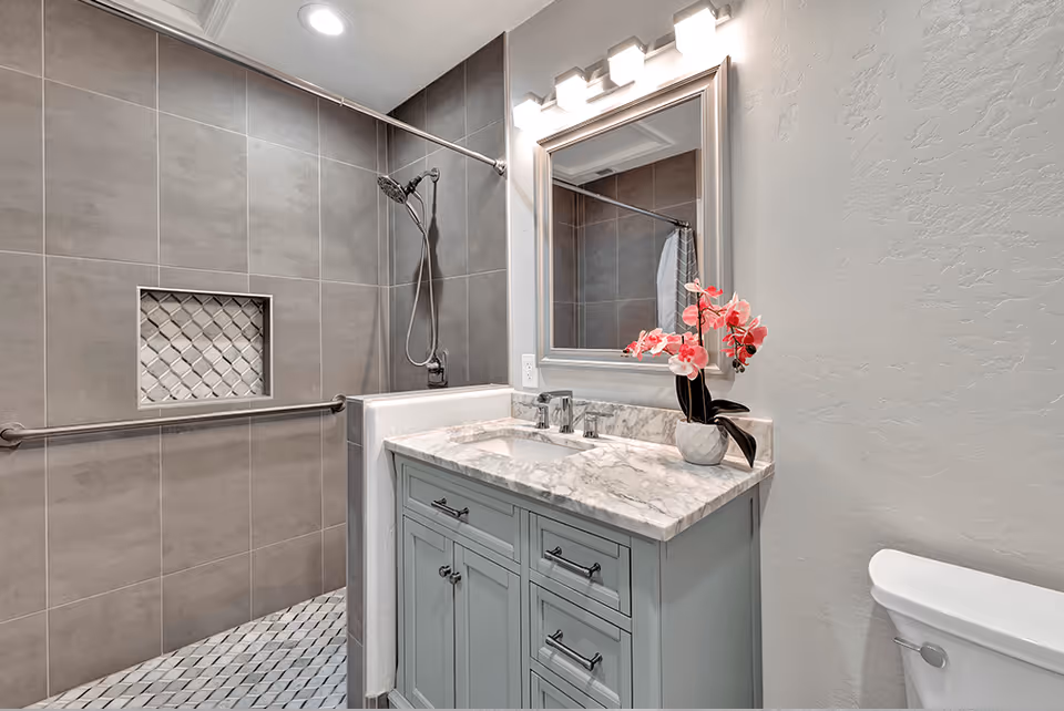 A modern bathroom featuring a walk-in shower with gray tiles and a built-in niche, a light blue vanity with a marble countertop, a rectangular mirror with lights above it, and a white toilet. A vase with pink flowers is placed on the vanity.