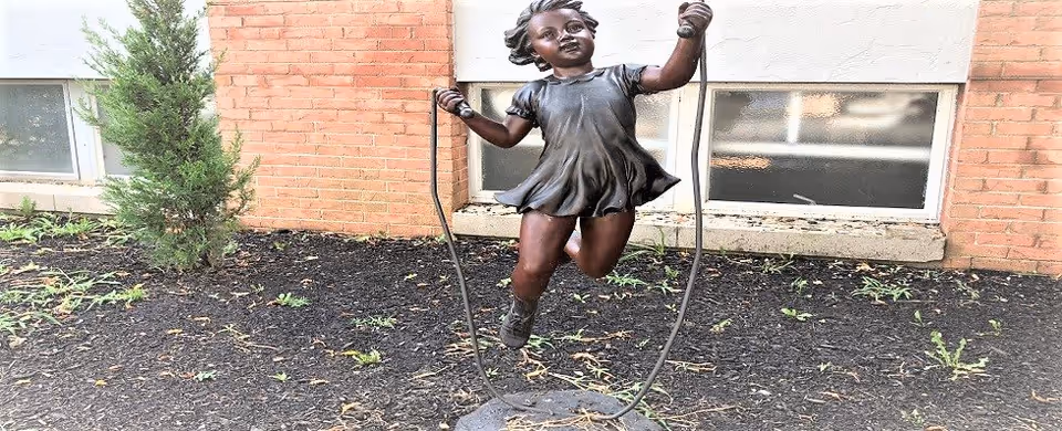 Bronze statue of a young girl skipping rope in a garden bed with mulch and small plants, set against a brick building with two windows.