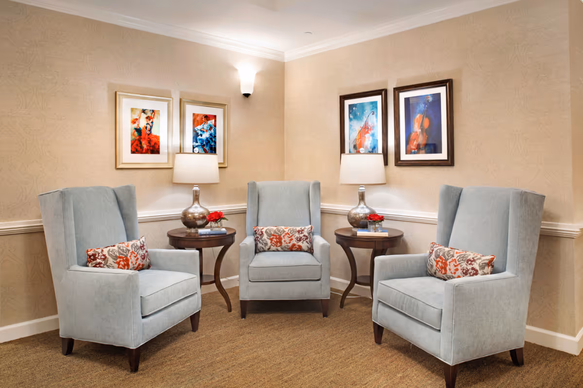 Three light-blue armchairs around side tables with lamps and colorful framed artwork on beige walls in a sitting area.