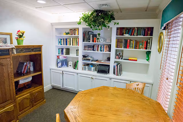 A cozy room with a wooden octagonal table and chairs in the foreground. In the background, there are white built-in bookshelves filled with books and decorative items, with a green plant hanging from the ceiling. To the left, there is a wooden cabinet with more books and a small potted plant with flowers on top. A window with wooden blinds is on the right side of the room.