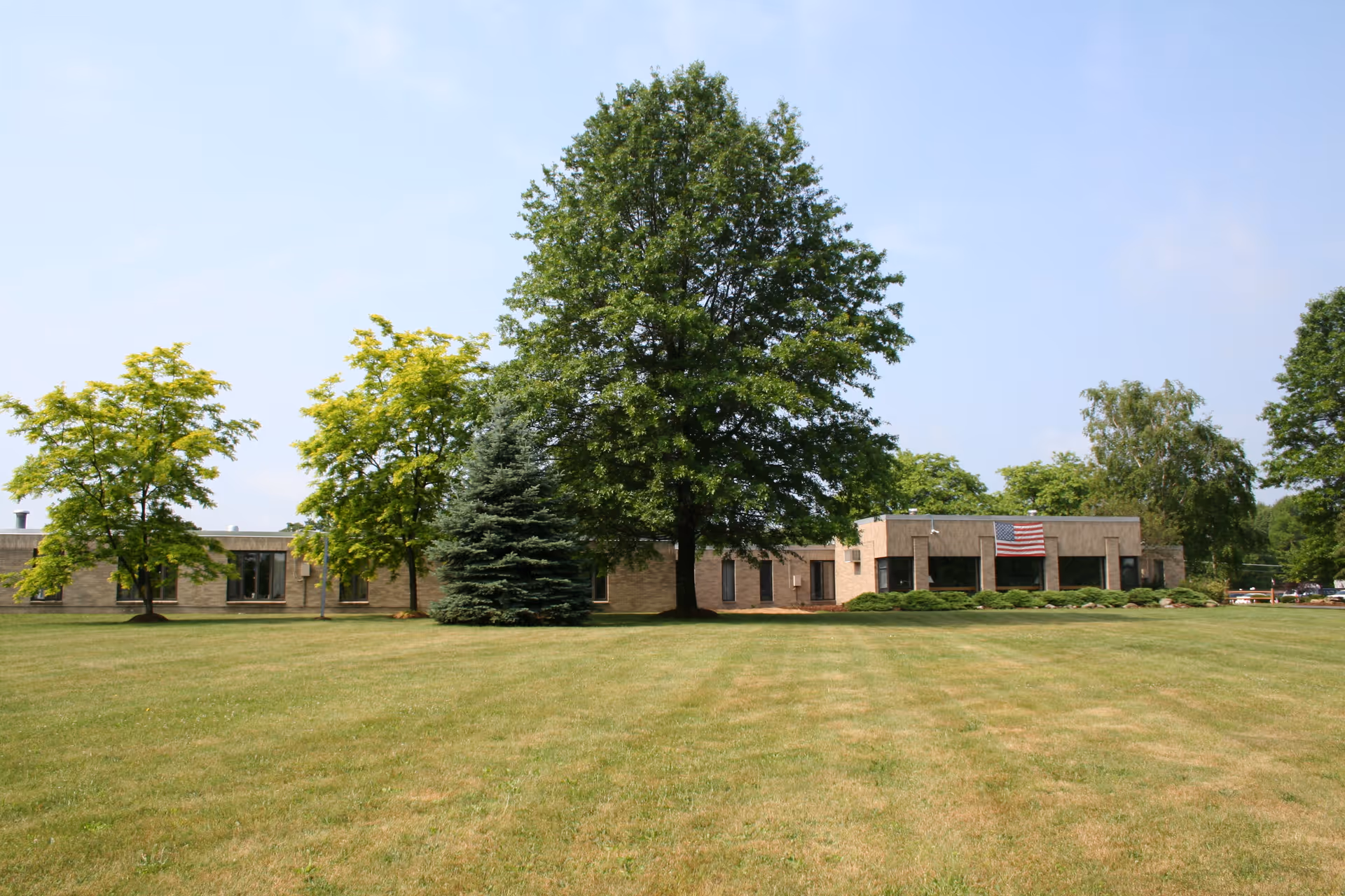 A single-story brick building with an American flag displayed on the front, surrounded by green trees and a large grassy lawn under a clear blue sky.