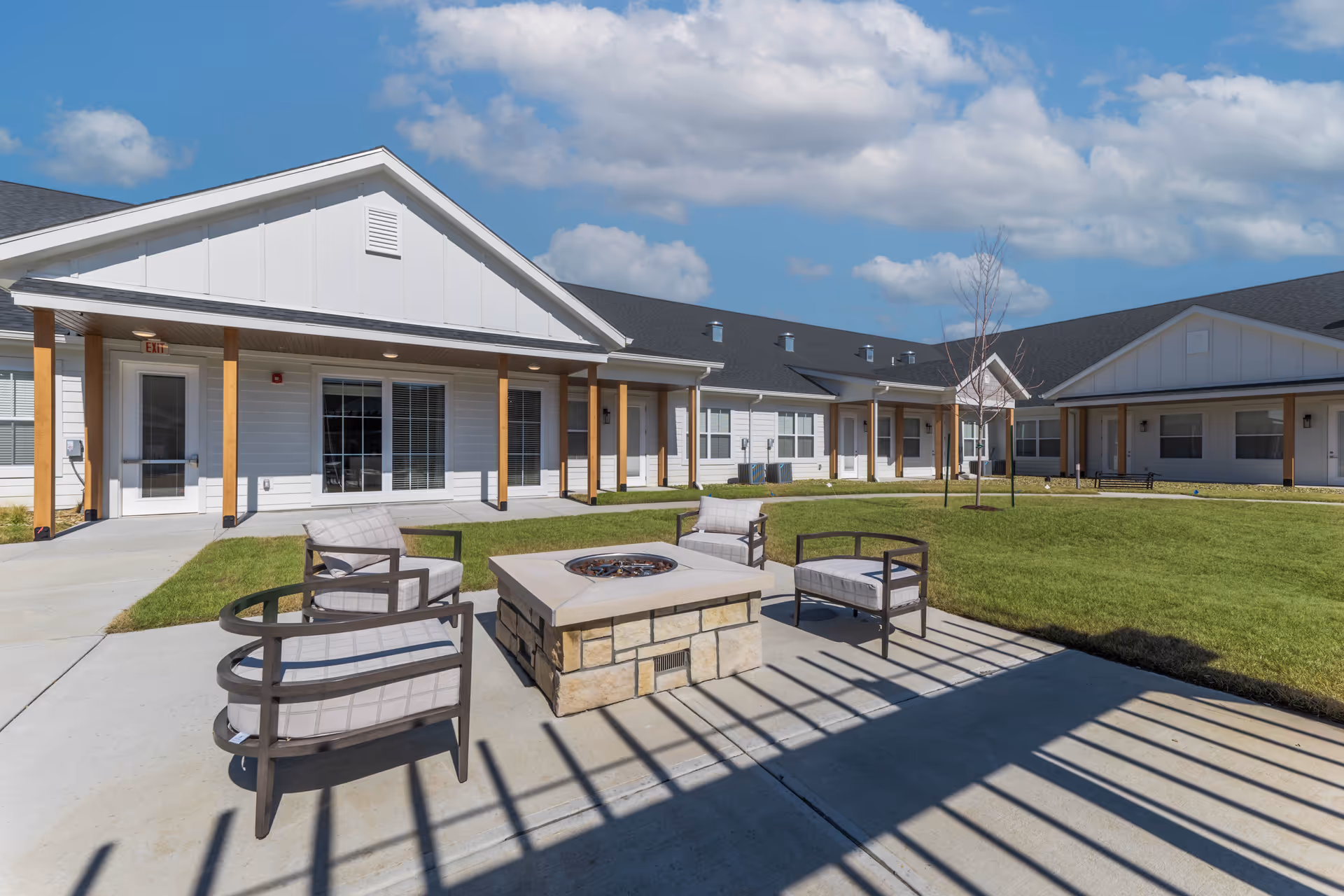 Outdoor courtyard with patio chairs arranged around a stone firepit and single-story white buildings surrounding a green lawn.