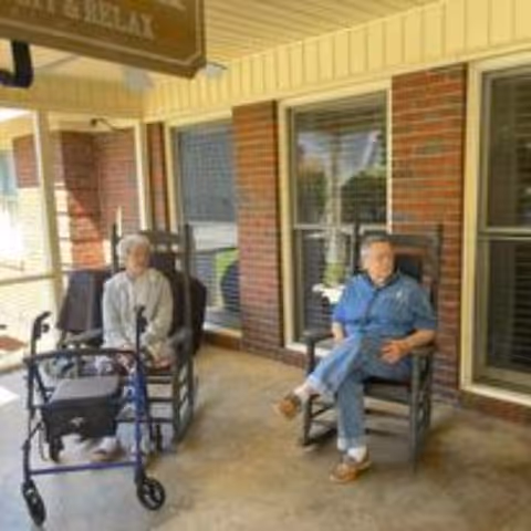Two elderly individuals sitting on rocking chairs on a covered porch outside a brick building. One person has a walker next to them. The porch has large windows and a sign above that partially reads 'RELAX'.