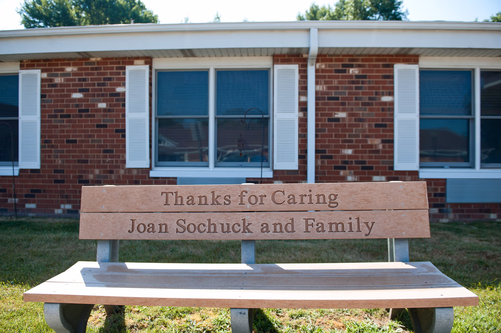 A wooden bench with the inscription 'Thanks for Caring Joan Sochuck and Family' in front of a brick building with white window shutters and blue windows.
