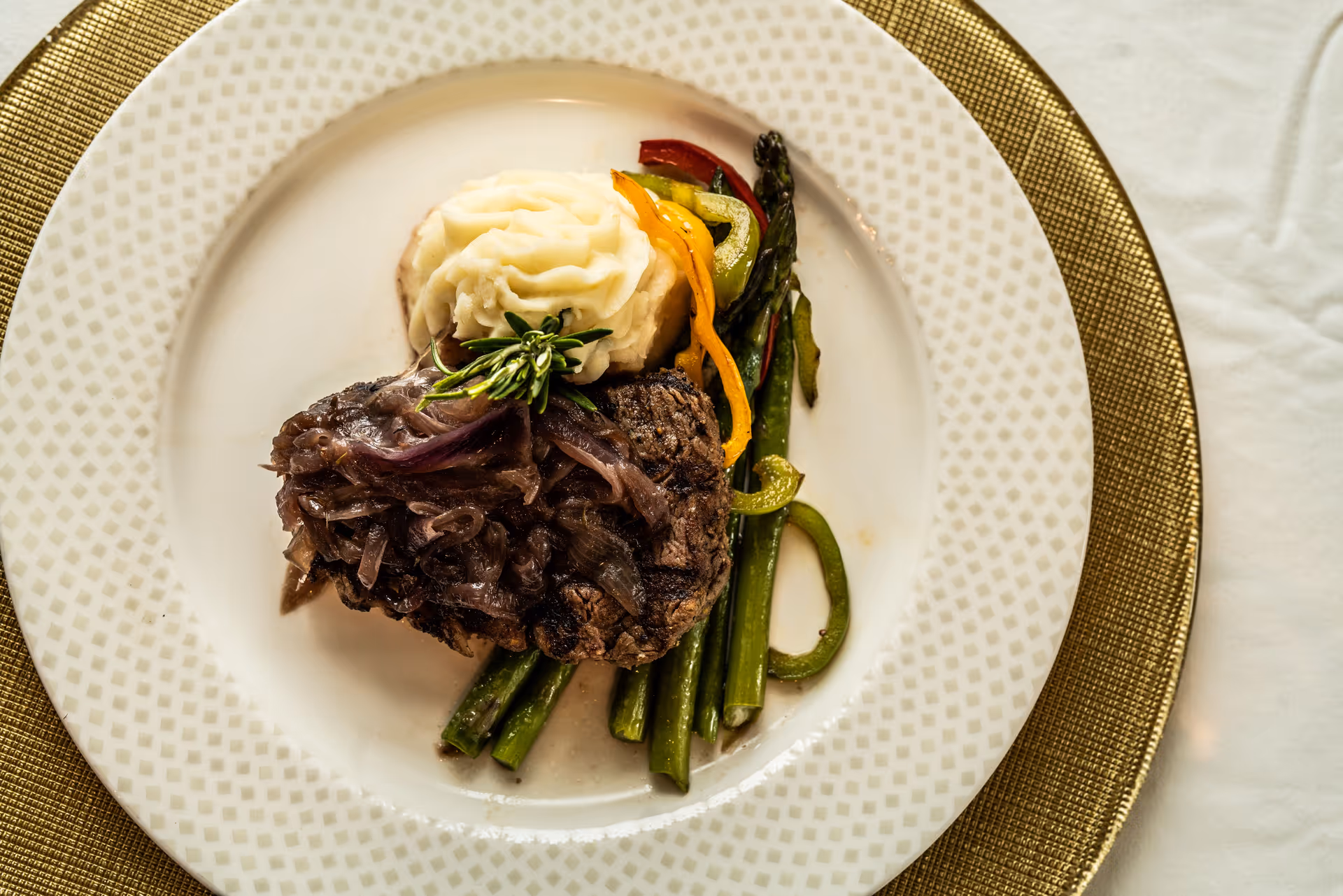 A plated meal consisting of a grilled steak topped with caramelized onions, served with mashed potatoes and a side of asparagus and sliced bell peppers on a white patterned plate with a gold charger underneath.