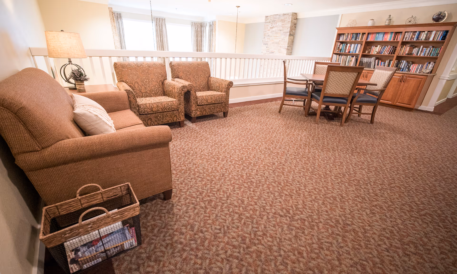 A cozy senior living common area with a brown upholstered sofa, two patterned armchairs, a wooden table with four chairs, a large wooden bookshelf filled with books, a lamp on a side table, and a basket with magazines on the floor. The room has carpeted flooring and large windows with curtains allowing natural light.