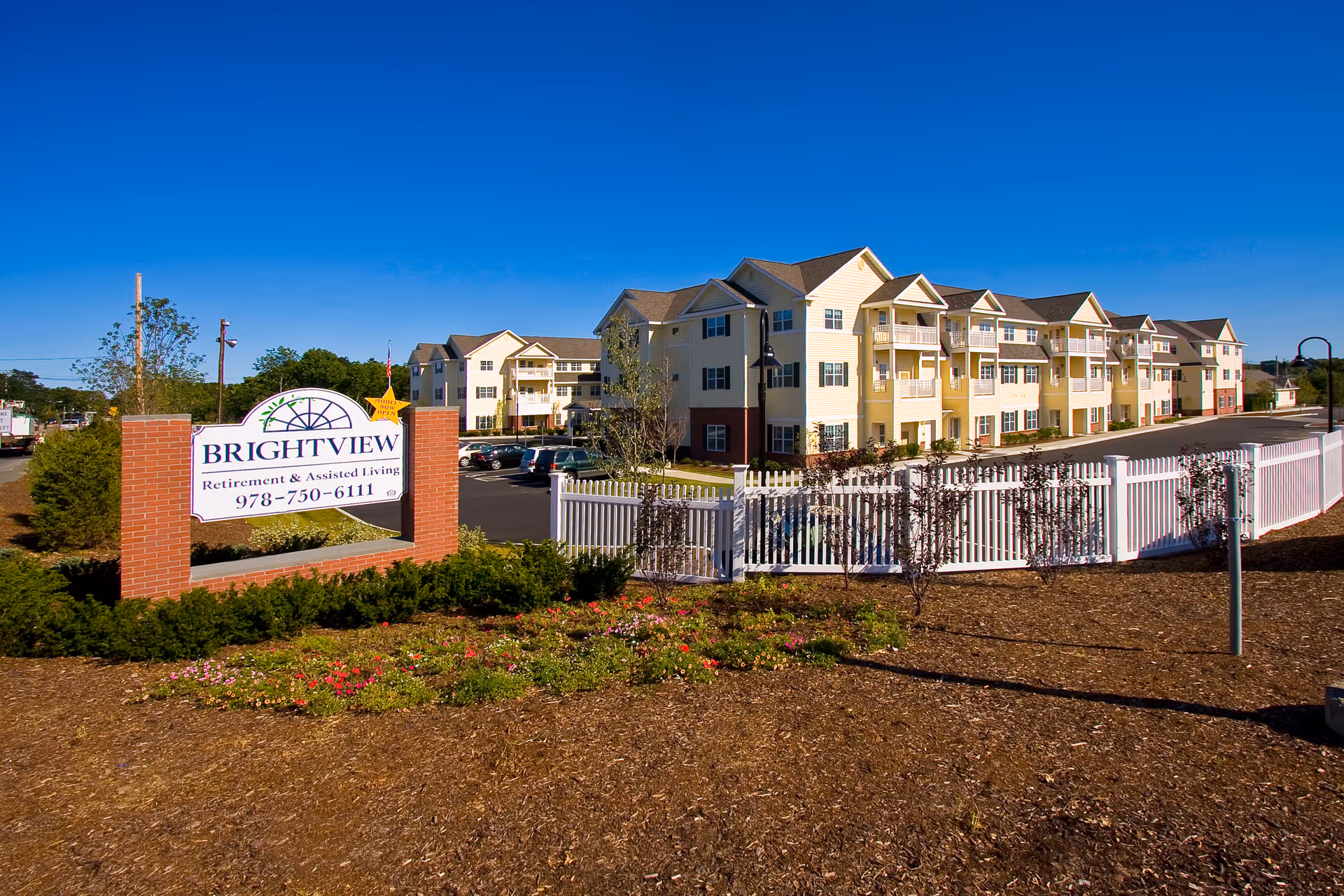 Exterior view of Brightview Danvers senior living facility showing a large multi-story building with balconies, a white fence, landscaped garden with flowers, and a clear blue sky. A sign in front reads 'Brightview Retirement & Assisted Living 978-750-6111'.
