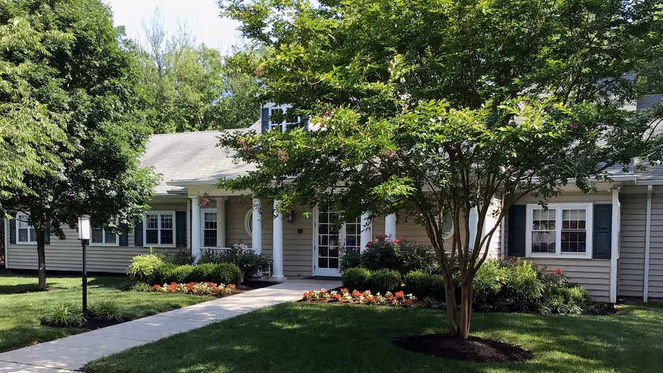 Exterior view of a single-story building with beige siding and white columns at the entrance. The building is surrounded by green trees, bushes, and a well-maintained lawn with a concrete walkway leading to the front door. Flower beds with colorful flowers line the walkway.
