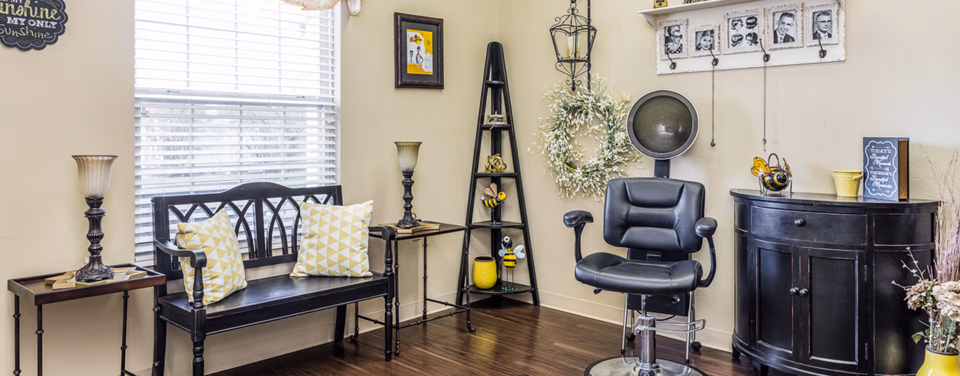 A cozy salon area with a black salon chair in front of a vintage hair dryer. To the left, there is a black wooden bench with two yellow and white patterned pillows, flanked by two small tables each holding a lamp. The walls are decorated with framed pictures, a wreath, and a shelf with hooks holding black and white photos. The floor is dark wood, and a large window with white blinds allows natural light to fill the room.