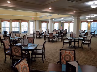 A spacious dining room with multiple dark wood tables and chairs featuring patterned cushions. Large windows with arched tops allow natural light to fill the room. The ceiling has recessed lighting and ceiling fans, and the carpet has a subtle patterned design.