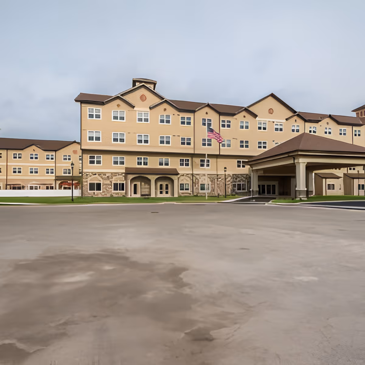 Exterior view of a multi-story senior living facility building with beige walls, multiple windows, and a covered entrance. An American flag is flying on a flagpole in front of the building.