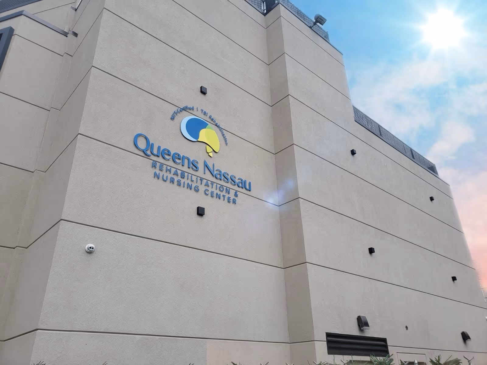 Exterior view of the beige multi-story facade of Queens Nassau Rehabilitation & Nursing Center with its logo and name mounted on the wall under a blue sky.
