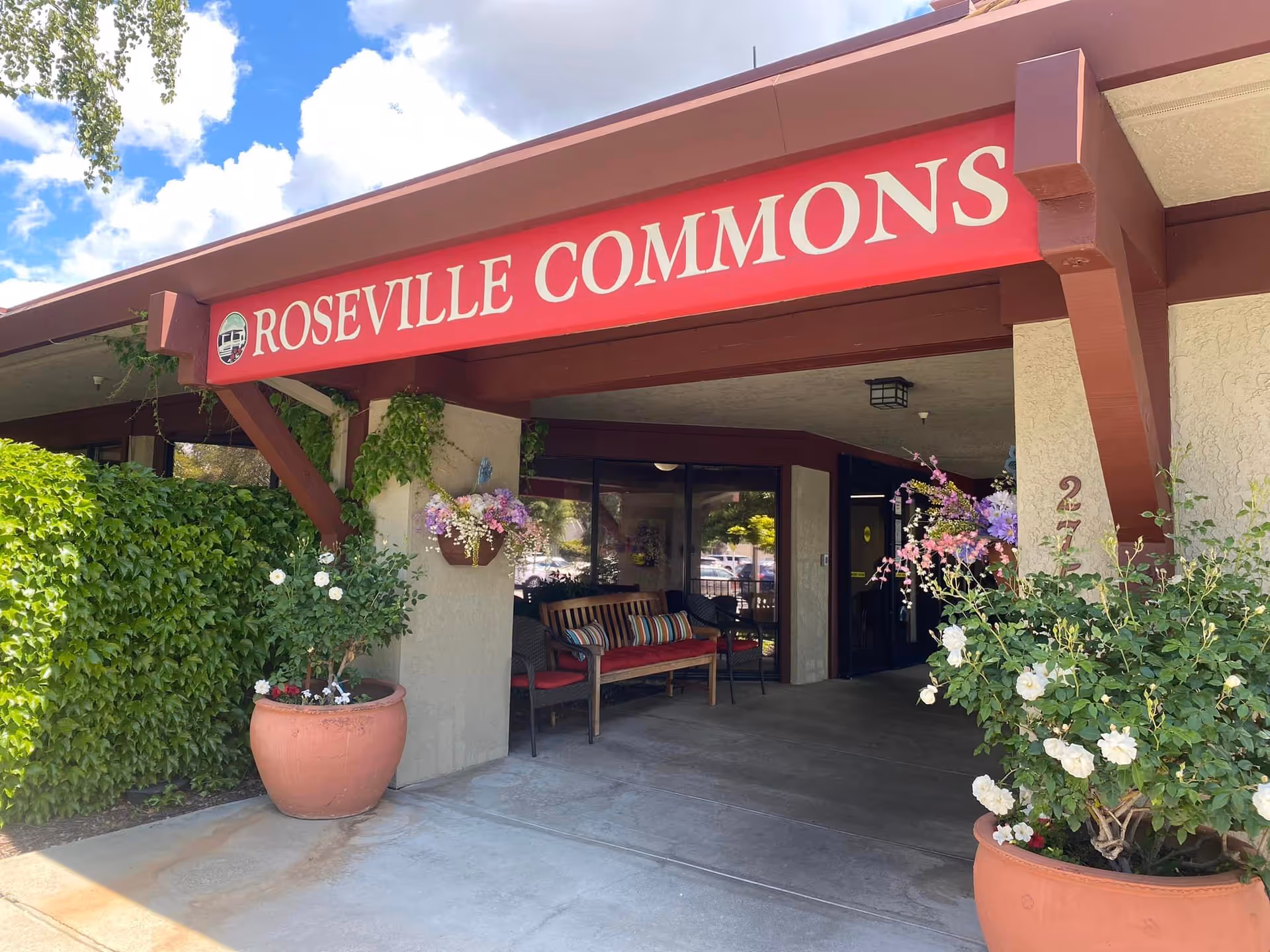 Front entrance of Roseville Commons senior living with a red sign, potted plants, and a bench under a covered entryway.