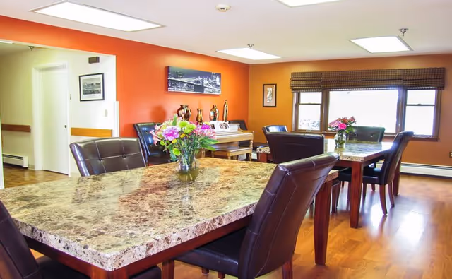 A bright dining room with two rectangular marble-patterned tables, each adorned with a vase of fresh flowers. The room has wooden flooring, leather cushioned chairs, a large window with blinds, and an orange accent wall featuring framed pictures and decorative items on a piano.
