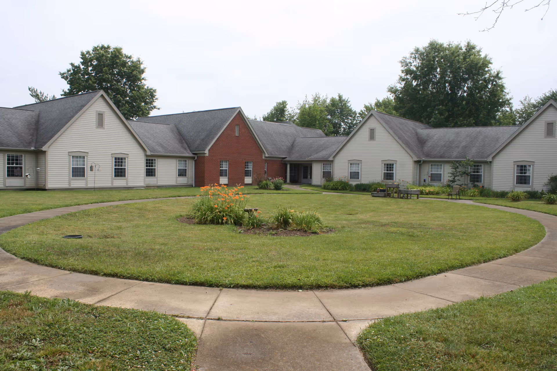 Single-story senior living facility building with beige siding and a red brick section in the center, surrounding a circular grassy courtyard with a flower bed in the middle and a concrete walkway around it. Trees are visible in the background under a cloudy sky.
