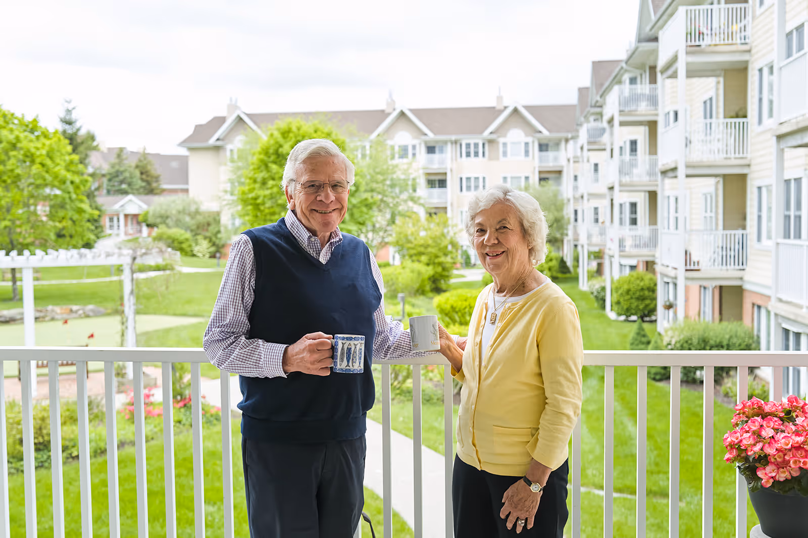 An elderly man and woman standing on a balcony holding coffee mugs, smiling at the camera. Behind them is a well-maintained garden area with green grass, trees, and a multi-story residential building.