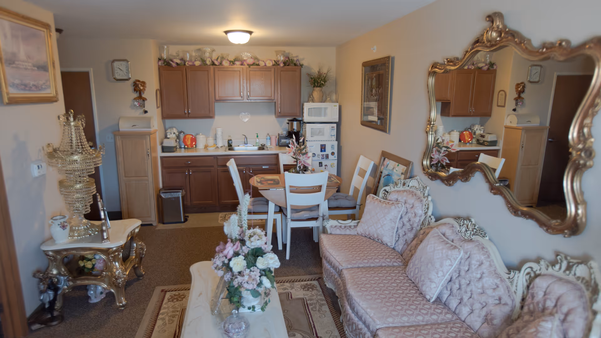 A cozy living area with a vintage-style sofa adorned with patterned cushions, a large ornate mirror on the wall, and a small dining table with four chairs. The background features a compact kitchen with wooden cabinets, a microwave, a refrigerator, and various kitchen items. Decorative flowers and a chandelier-style lamp add to the room's charm.