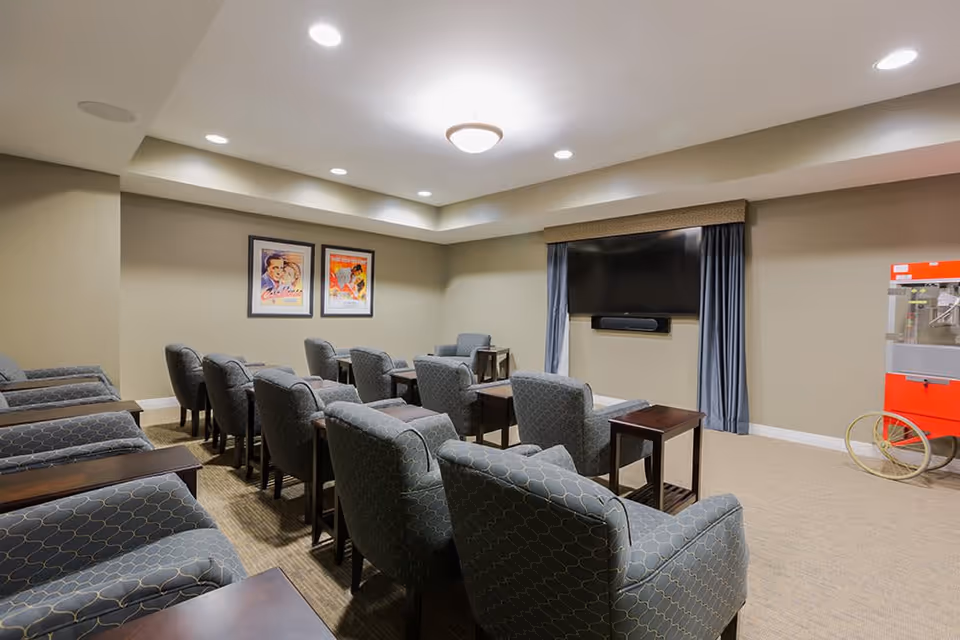 A cozy movie or media room with multiple rows of cushioned armchairs facing a large flat-screen TV mounted on the wall. The room has beige walls, recessed lighting, two framed vintage-style posters on one wall, and a red popcorn machine on wheels in the corner.
