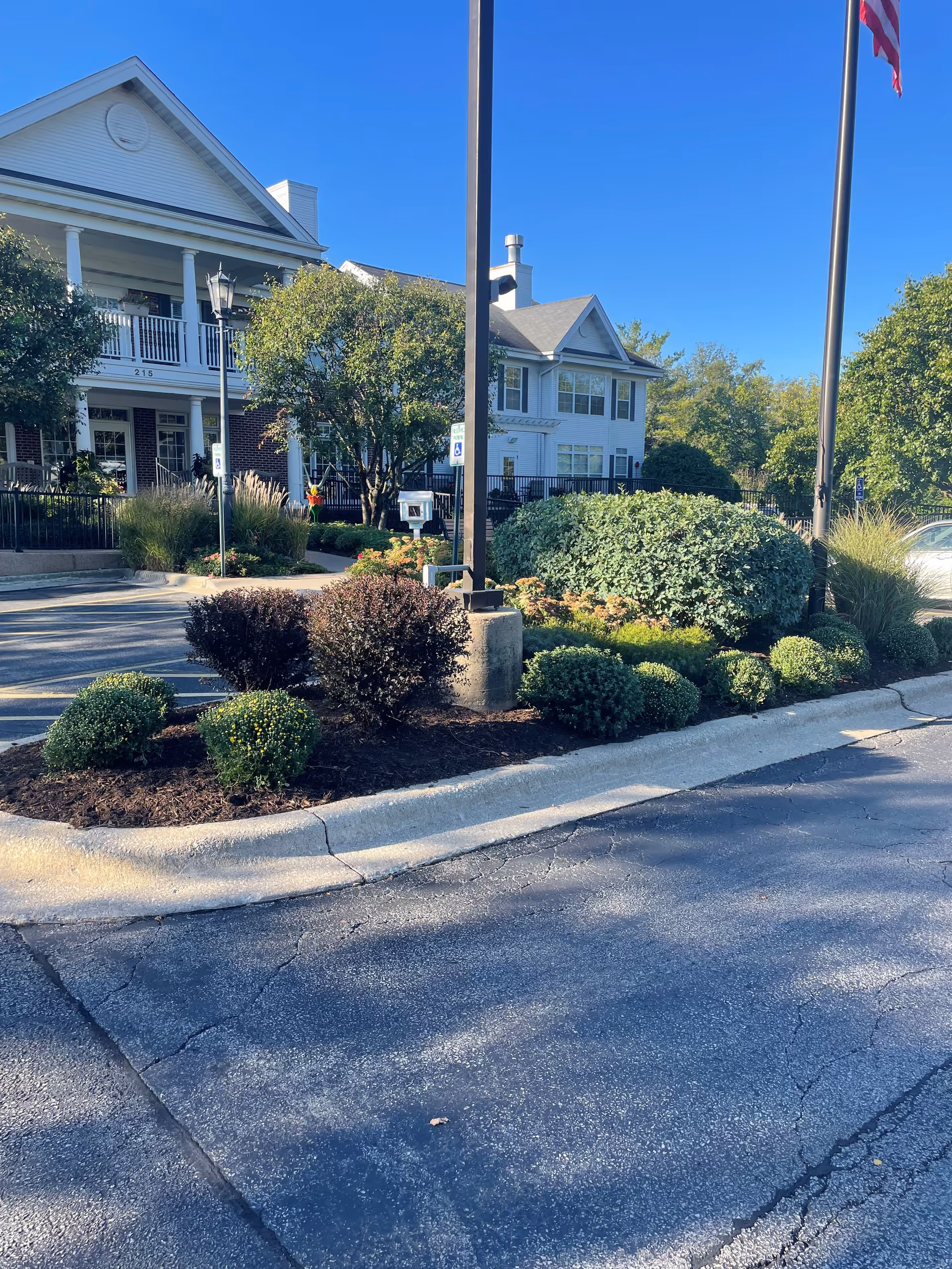 Front exterior of a multi-story assisted living building with landscaped shrubs, a flagpole, and a parking area.