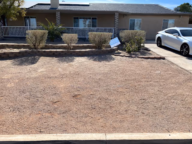 Single-story building front with a gravel yard, low shrubs, a white porch railing, and a white car parked at the driveway on the right.