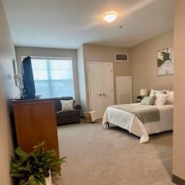 Bright neutral bedroom with a made bed in white and green linens, a wooden dresser with a TV, an armchair by a large window, and a potted plant.