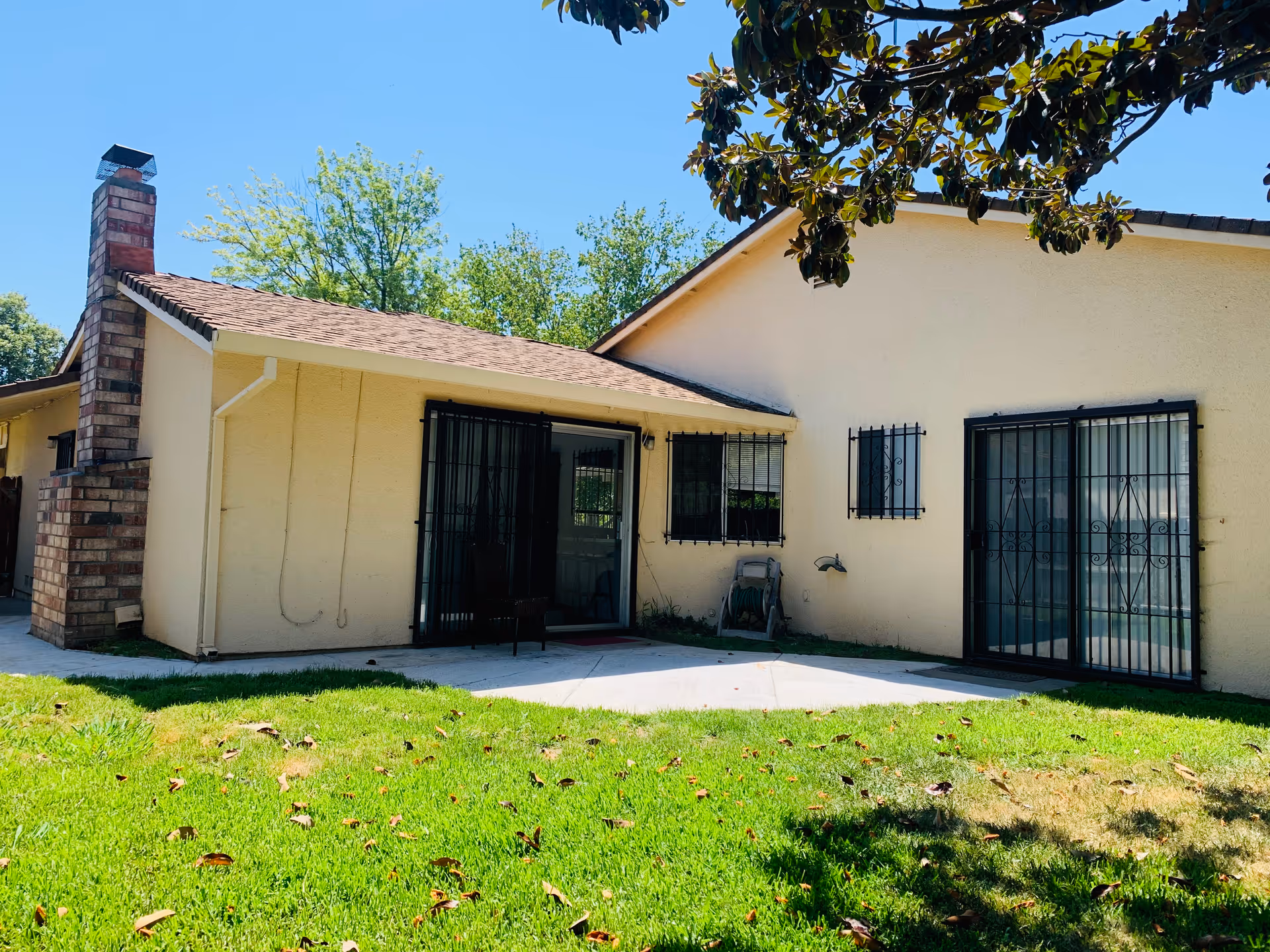 Exterior view of a single-story building with beige walls and a brown shingled roof. The building has black metal security bars on the windows and sliding glass doors. There is a brick chimney on the left side and a green lawn in the foreground with some fallen leaves. A tree branch with green leaves is visible at the top right corner.