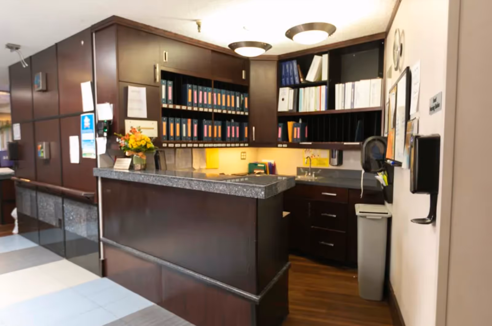 Reception or nurse station area with dark wood cabinetry and shelves filled with binders and files. A countertop with a flower arrangement and various papers is visible. The area is well-lit with ceiling lights and has a trash bin and hand sanitizer dispenser on the wall.