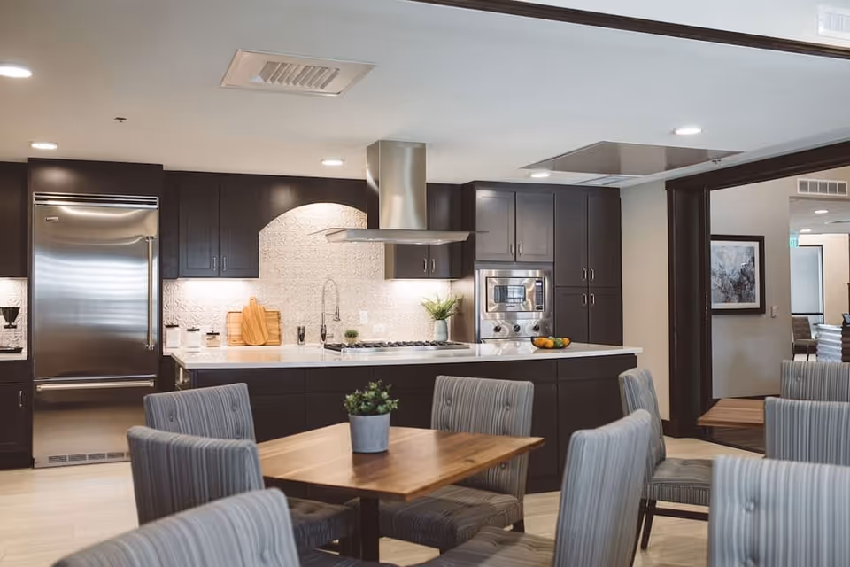 Communal dining area with wooden tables and upholstered chairs in front of a modern open kitchen with dark cabinets and stainless appliances.