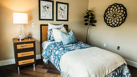 A neatly made single bed with blue floral bedding and multiple pillows in a warmly lit room. Next to the bed is a wooden nightstand with three drawers, a glass lamp, and decorative items. Two framed bird illustrations hang above the bed, and a decorative circular wall piece is mounted on the adjacent wall. A potted plant stands in the corner.