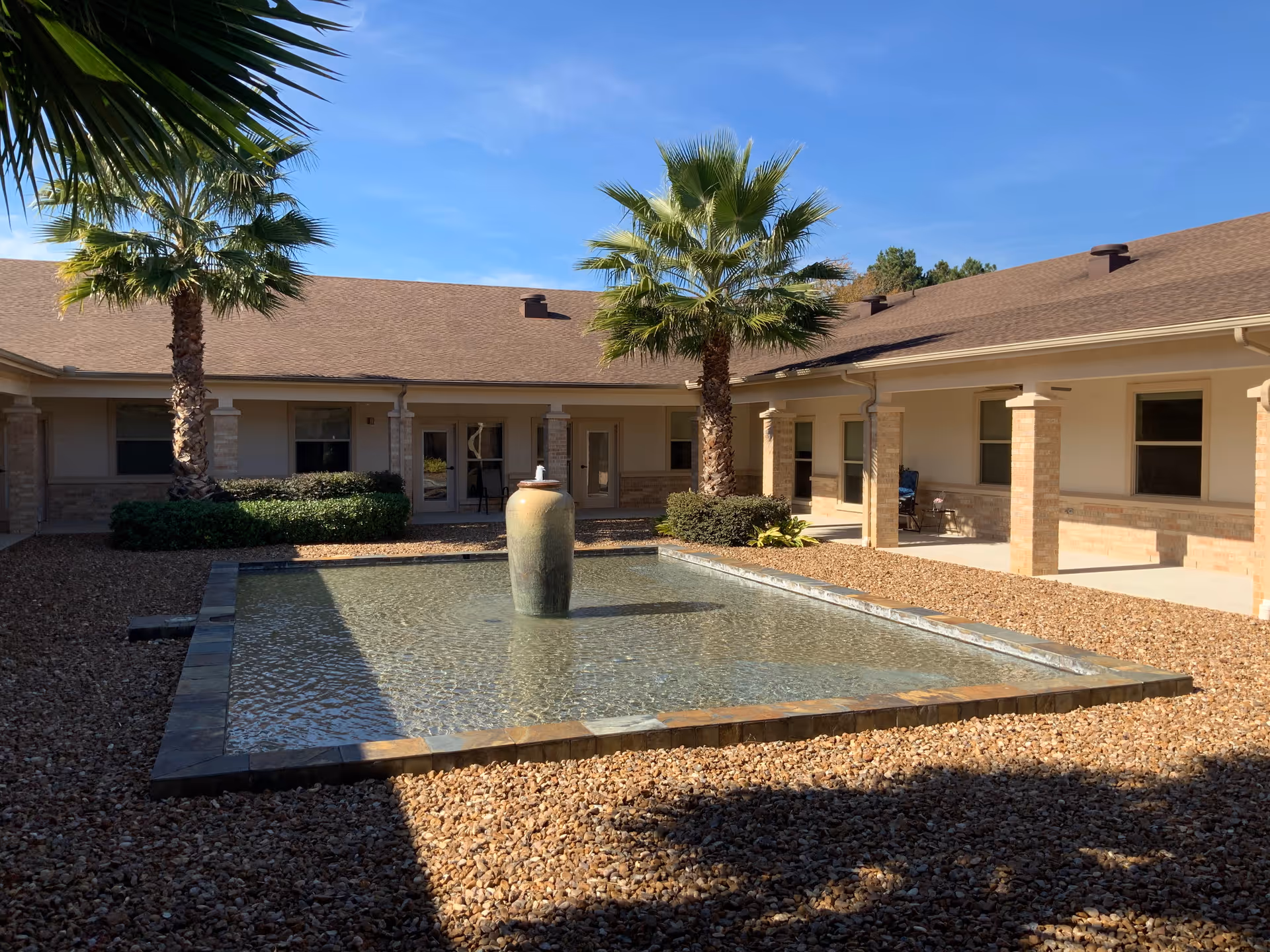 Courtyard with a shallow reflecting pool and central fountain, flanked by palm trees and a single-story building.