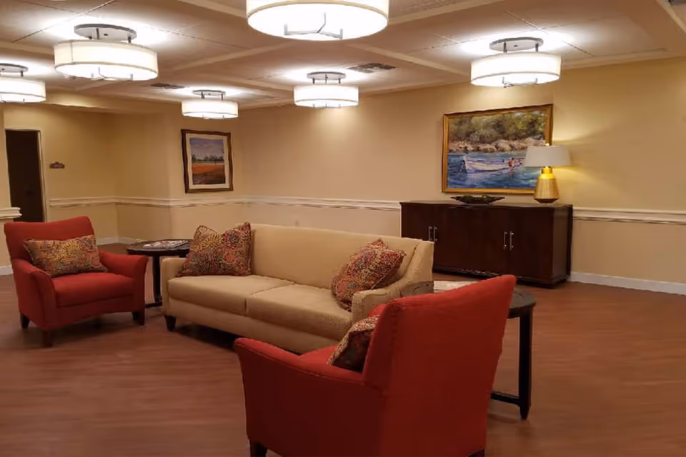 A cozy living room area with a beige sofa and two red armchairs, each with patterned cushions. The room has warm yellow walls, wooden flooring, and several round ceiling lights. There is a dark wooden sideboard against the wall with a lamp and a painting above it, as well as another framed picture on the adjacent wall.