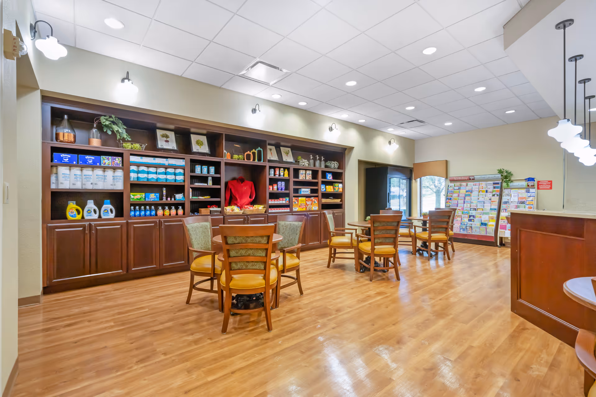 A well-lit room with wooden flooring featuring several round tables with chairs arranged around them. Along one wall, there are built-in wooden shelves stocked with various household and personal care products. A rack filled with brochures or pamphlets is visible near a window with blinds. The ceiling has recessed lighting and hanging pendant lights.