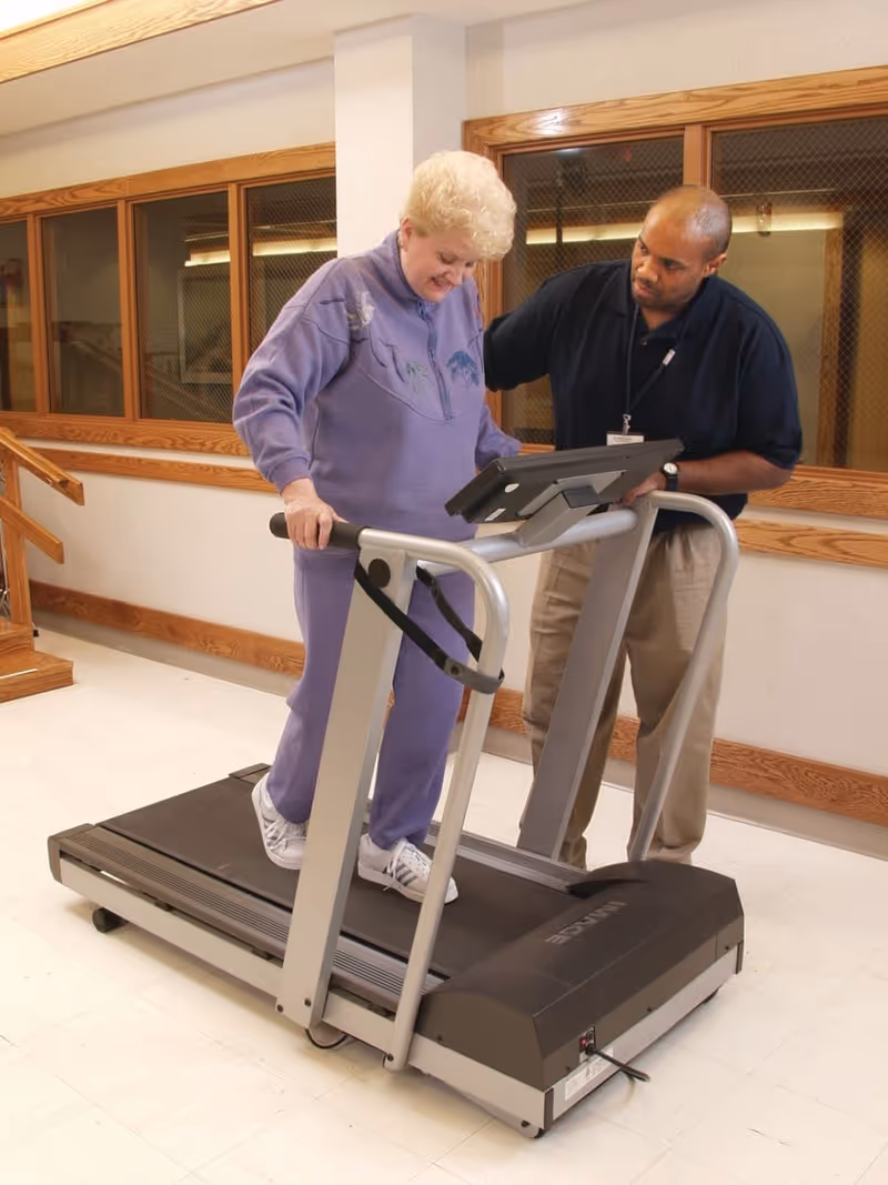 An elderly woman in a purple tracksuit is walking on a treadmill while a man in a navy blue shirt and beige pants stands beside her, offering support and guidance. They are indoors in a room with wooden window frames and a light-colored floor.