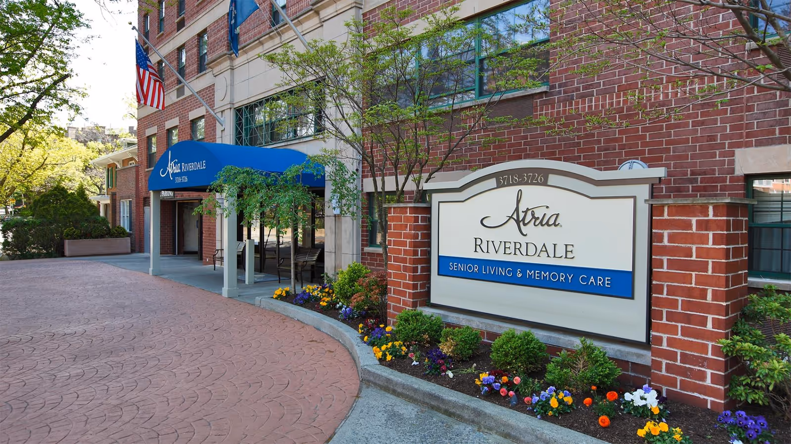 Entrance and sign for Atria Riverdale senior living and memory care with a blue awning, American flag, and landscaped flowerbed.