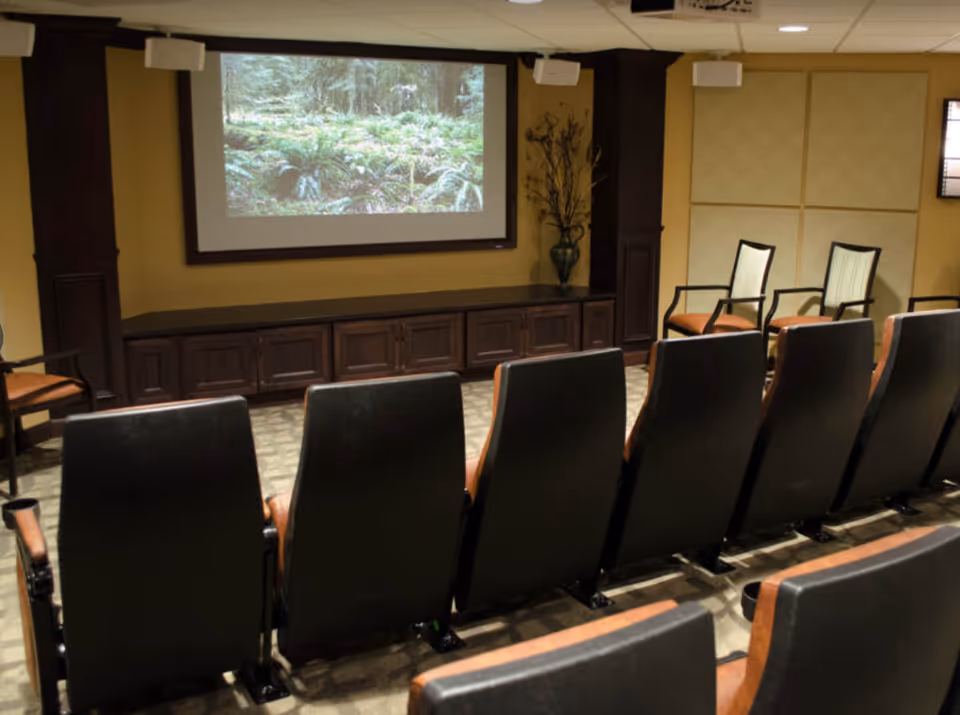 A small theater room with two rows of black and brown cushioned chairs facing a large screen displaying an image of a forest. The room has beige walls, dark wood paneling, and additional chairs along the side wall.
