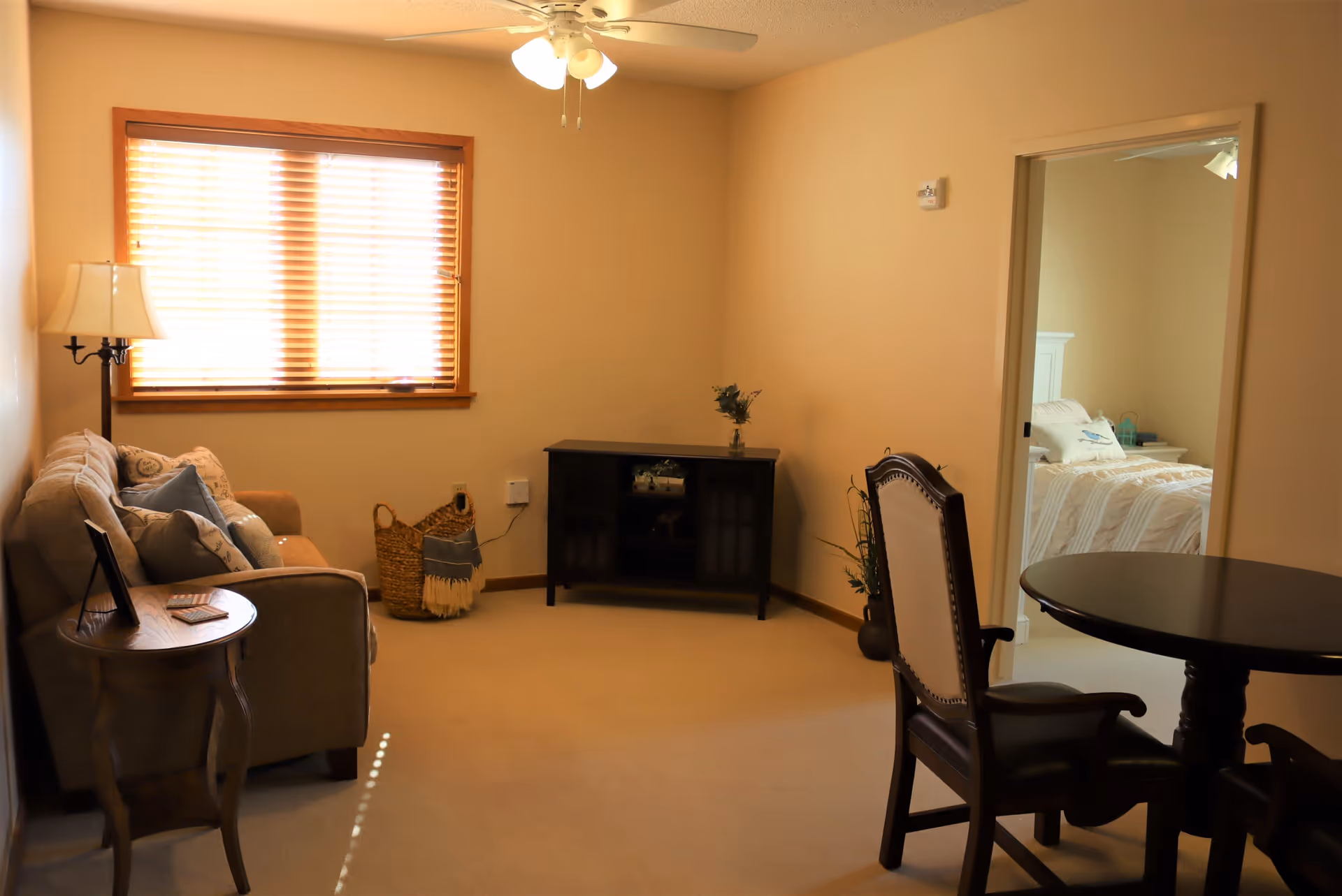 A cozy living room with a beige couch adorned with several pillows, a wooden side table with a lamp, a dark wooden cabinet against the wall, and a round wooden table with two chairs. A window with wooden blinds lets in natural light. An open doorway reveals a bedroom with a white bed and pillows.