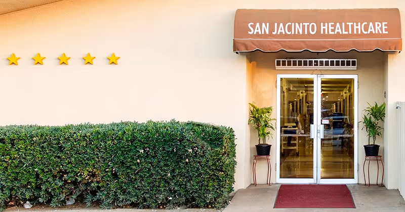 Entrance to San Jacinto Healthcare facility with a brown awning displaying the facility name, glass double doors, two potted plants on either side of the entrance, a red doormat, and a neatly trimmed hedge to the left with five gold stars mounted on the wall above it.