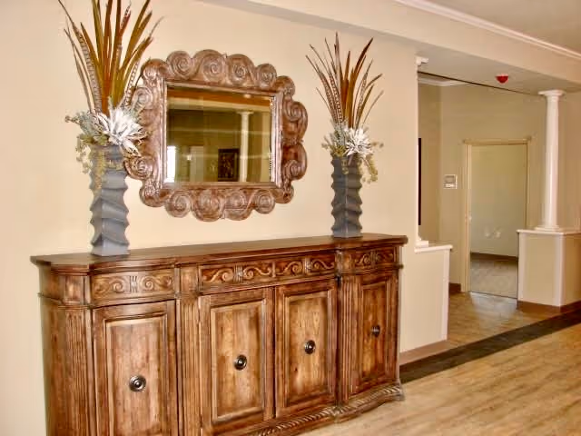 A hallway area featuring a wooden sideboard with ornate carvings and four cabinet doors. On top of the sideboard are two tall vases with decorative dried plants. Above the sideboard hangs a large, intricately carved wooden framed mirror. The walls are painted beige, and the floor has a light wood finish. In the background, there is an open doorway and a white column supporting a partial wall.