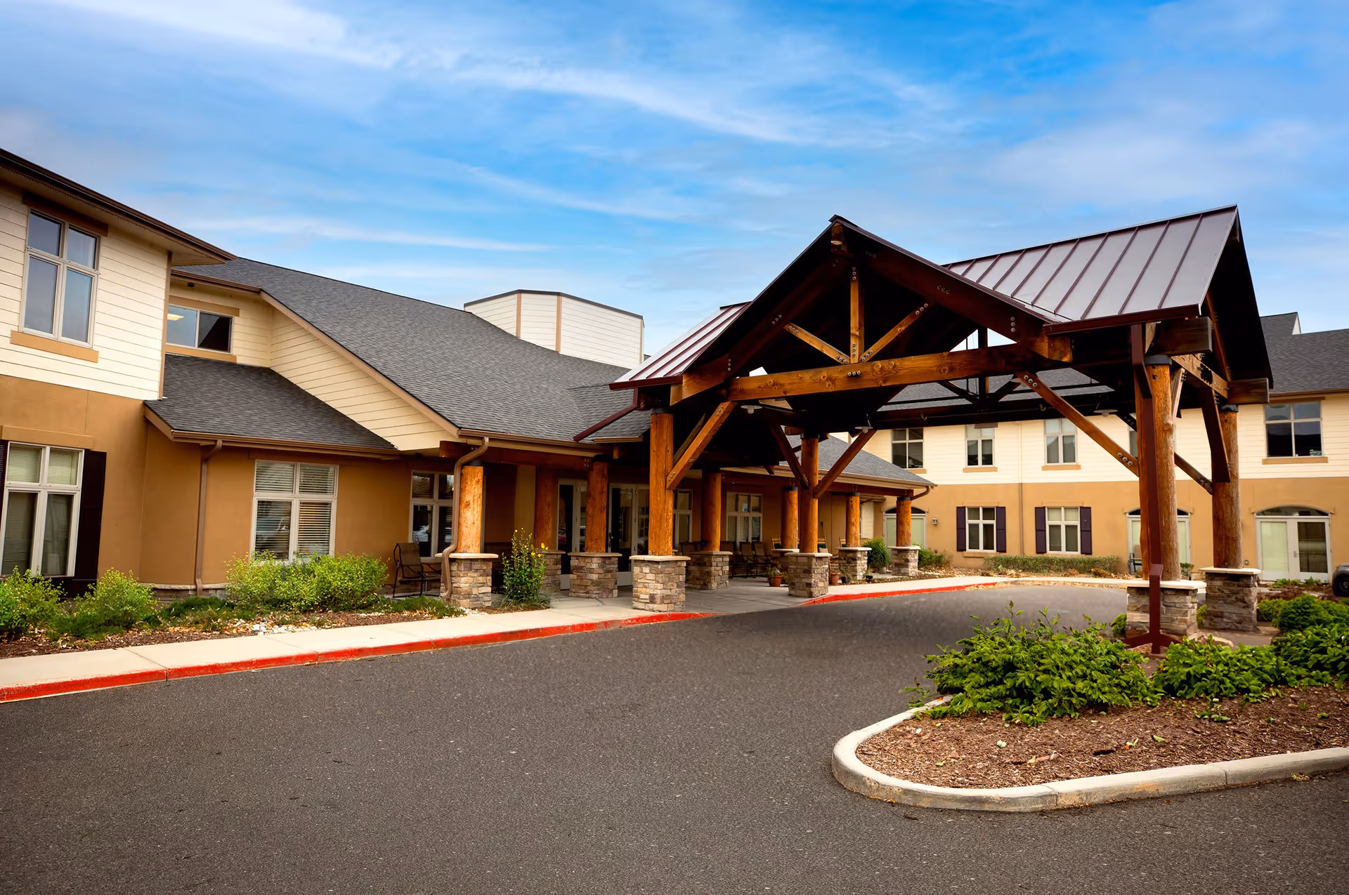 Exterior view of MeadowView of Greeley senior living facility showing a covered entrance with wooden beams and stone pillars, surrounded by a paved driveway and landscaped greenery under a blue sky.