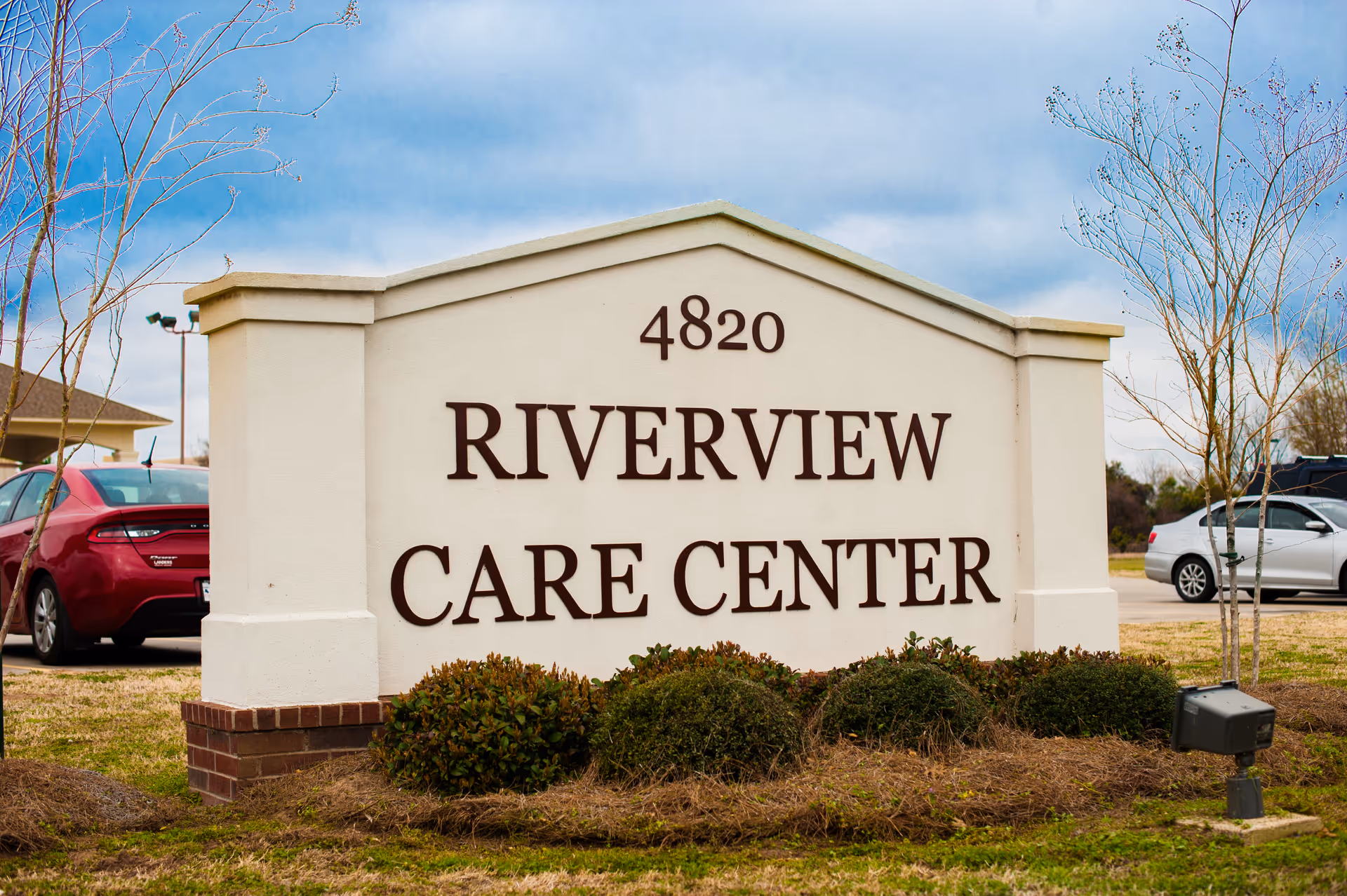 Outdoor view of a large white sign with the address number 4820 and the name Riverview Care Center, surrounded by small bushes and grass, with parked cars and a cloudy sky in the background.