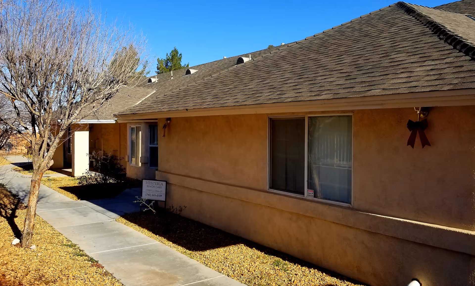Exterior view of a single-story senior care facility building with a beige stucco finish and a shingled roof. There is a sidewalk running alongside the building, a leafless tree, and a small sign near the entrance that reads 'Hesperia Senior Care'. The sky is clear and blue.